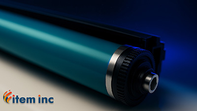 Close-up macro shot of a laser-printer drum unit featuring a glossy teal photoconductor cylinder, metal end cap and black gear in sharp focus on a reflective white surface, bathed in deep navy blue (#094670) rim lighting; an orange-and-yellow swirling “item inc” logo appears in the lower-left corner.
