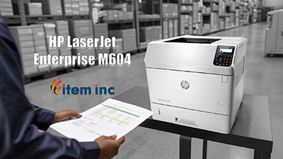 In a warehouse aisle, an HP LaserJet Enterprise M604 printer sits on a black table while a technician in the foreground holds a freshly printed report; overlaid text reads ‘HP LaserJet Enterprise M604’ next to the Item Inc logo.