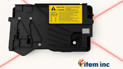 Close-up view of a laser/scanner assembly unit for a laser printer, shown against a white background with red laser beams crossing diagonally. A caution label is visible, and the Item Inc. logo appears in the corner.