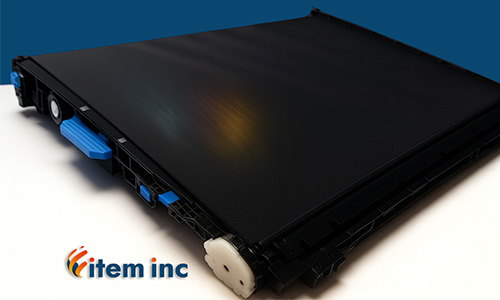 Close-up image of a printer transfer belt assembly on a white surface with a deep blue background. The black belt shows a subtle reflective CMYK gradient, and features include blue plastic levers and a white gear. The "item inc" logo is placed in the bottom left corner of the image.