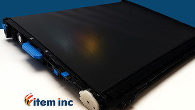 Close-up image of a printer transfer belt assembly on a white surface with a deep blue background. The black belt shows a subtle reflective CMYK gradient, and features include blue plastic levers and a white gear. The "item inc" logo is placed in the bottom left corner of the image.