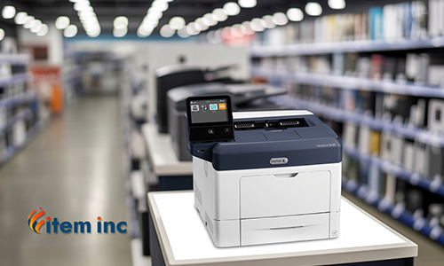A Xerox VersaLink B400 printer displayed on a store counter inside an office-equipment aisle, with shelves of products blurred in the background and the Item Inc logo visible in the foreground.