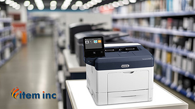 A Xerox VersaLink B400 printer displayed on a store counter inside an office-equipment aisle, with shelves of products blurred in the background and the Item Inc logo visible in the foreground.