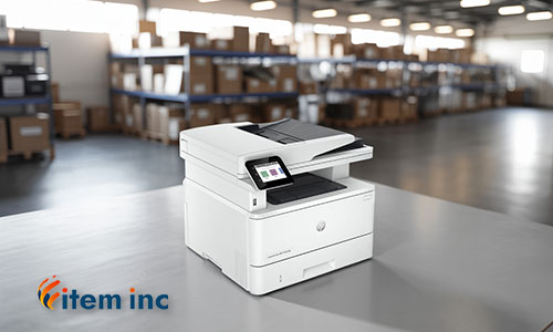 A white HP multifunction laser printer with a touchscreen display is sitting on a metal table. The touchscreen shows several colored app icons. The printer has an automatic document feeder on top and output trays below. The HP logo is visible on the front of the printer. In the bottom left corner, there is a logo for "item inc". The background is a blurred warehouse setting with shelves filled with cardboard boxes and bright lights on the ceiling.